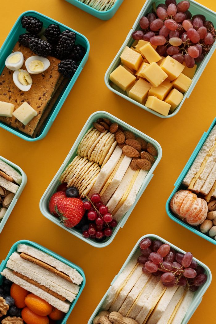 about-us-02 Assorted lunch boxes with snacks, fruits, and cheese on a vibrant yellow background.